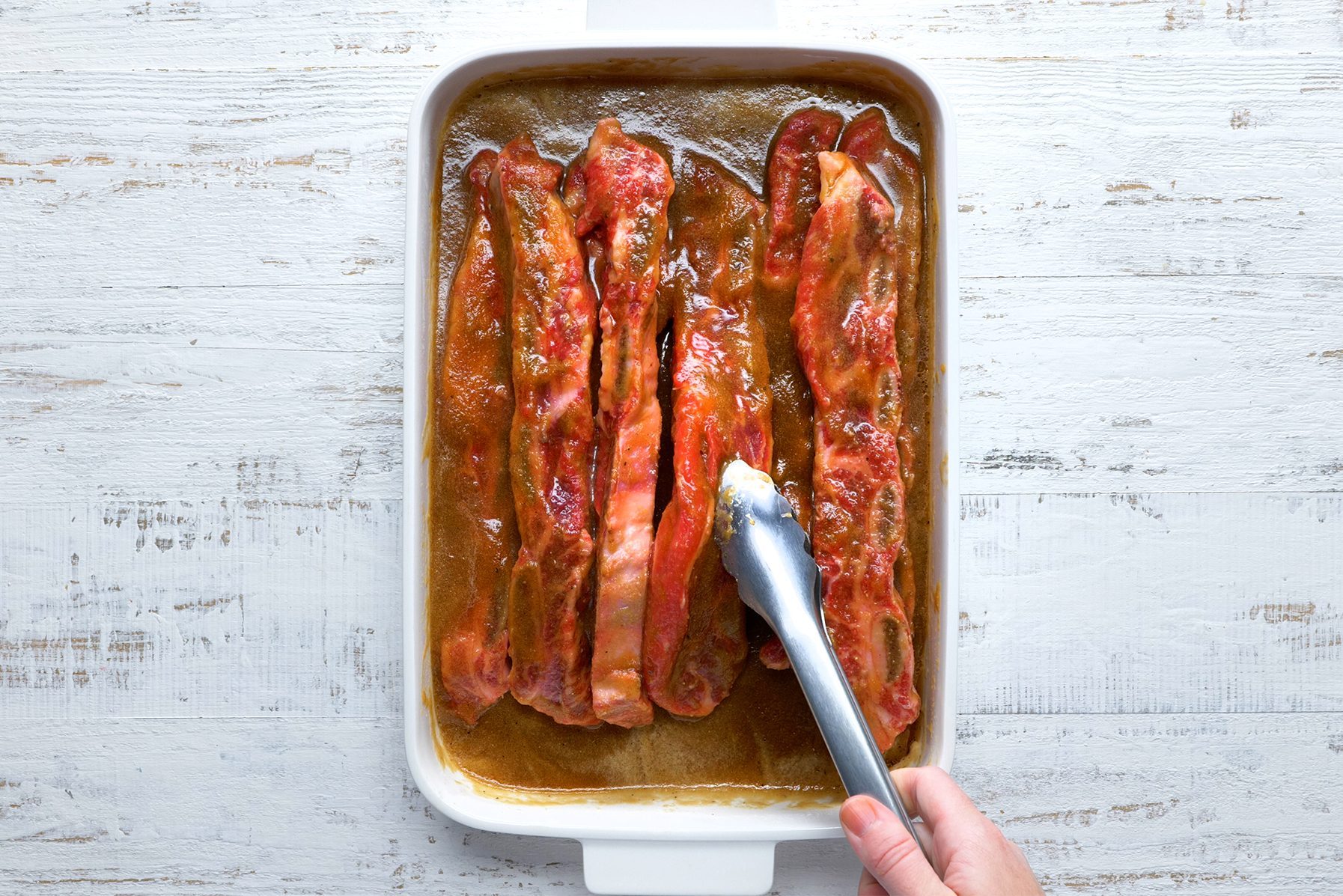 A hand uses tongs to place ribs in a white baking dish with a brown sauce. The dish is on a rustic white wooden surface.