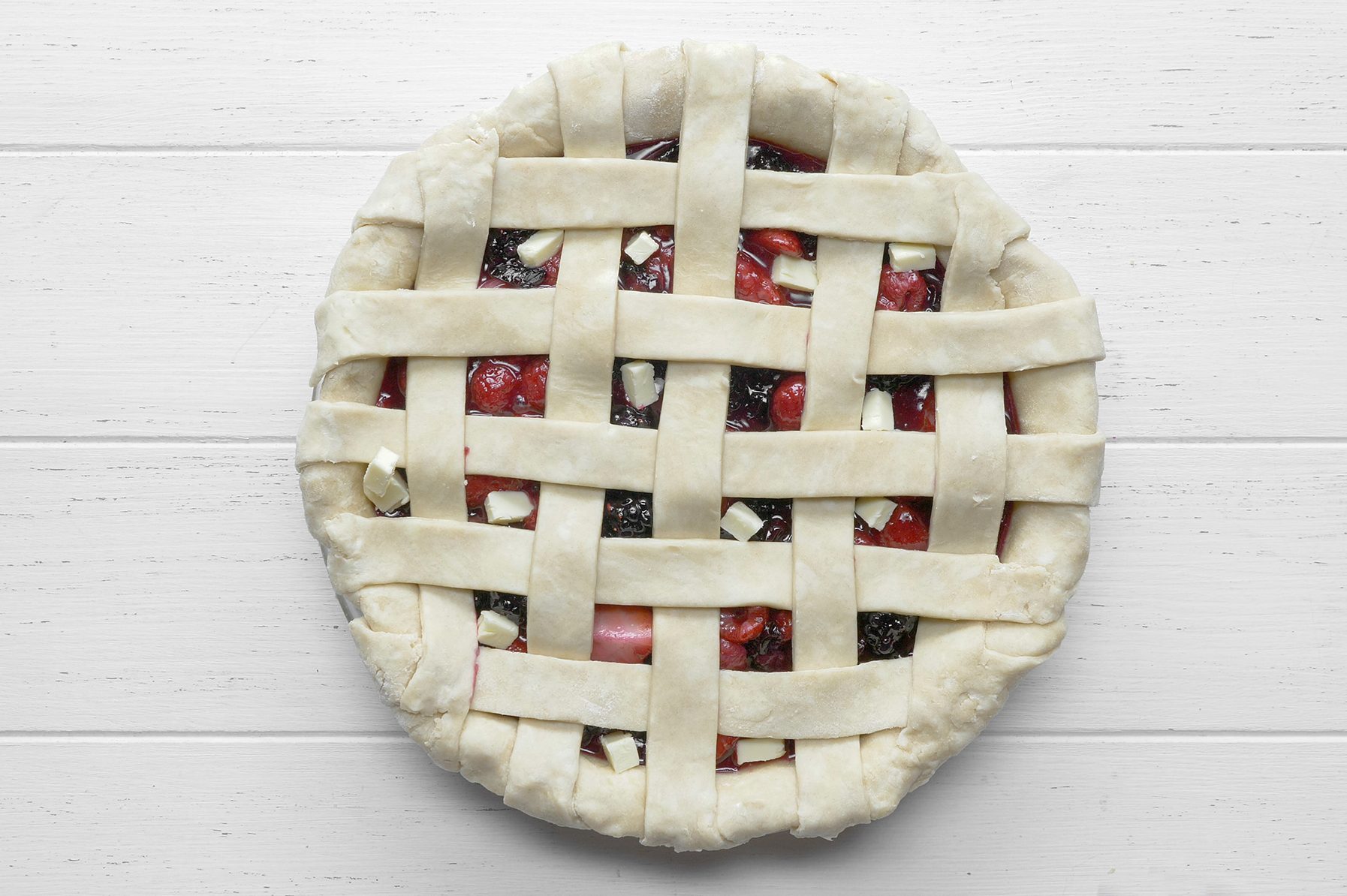 A top-down view of a lattice-topped cherry and berry pie on a white wooden surface. The golden-brown crust forms a woven pattern, revealing the red and dark purple fruits underneath.