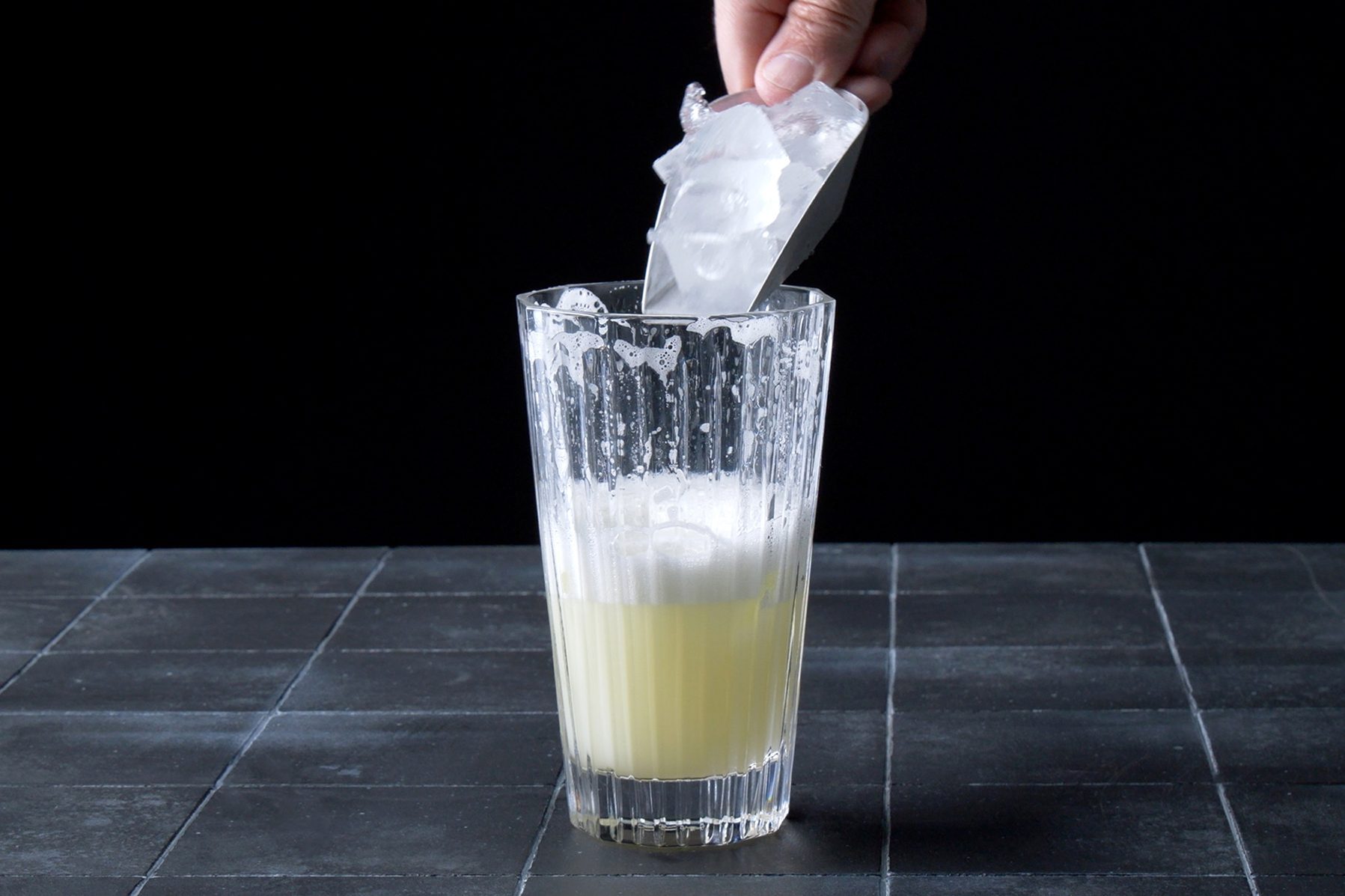A hand is adding ice cubes to a tall clear glass filled with a light yellow foamy liquid, set against a dark background on a tiled surface.