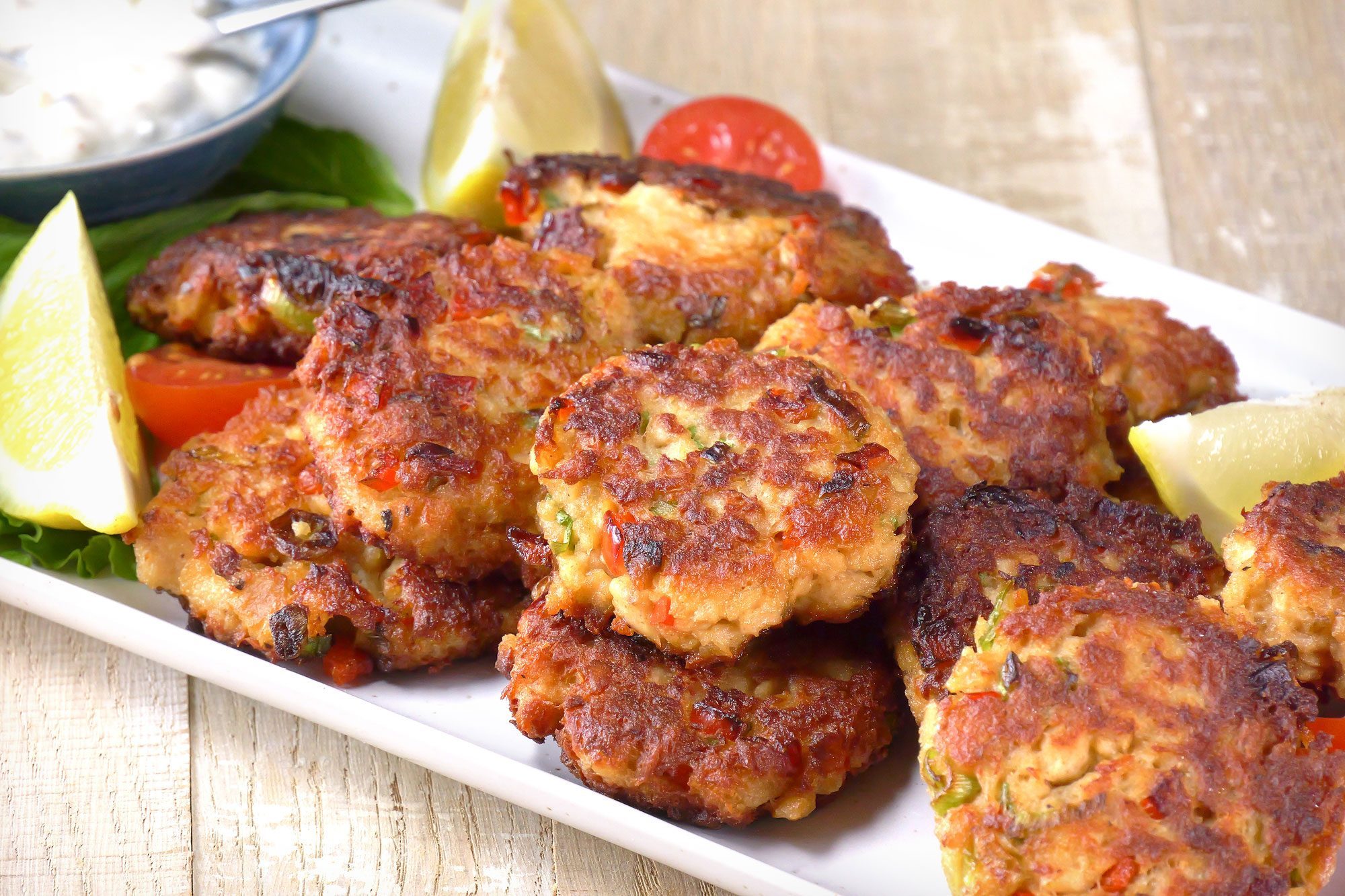 3/4 angle view shot of Tasty Salmon Croquettes; serve on large platter; with tartar sauce and lemon wedges; spoon; cherry tomatoes; wooden background;