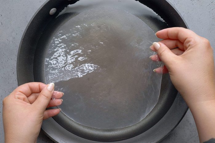 A person is holding a circular sheet of translucent rice paper with both hands above a shallow dish or pan filled with water.