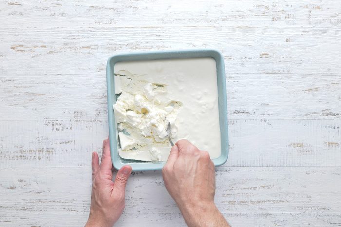 A person’s hands are seen holding a spoon and cutting into a square pan filled with a creamy white mixture, likely a dessert or dairy product, positioned on a white wooden surface.