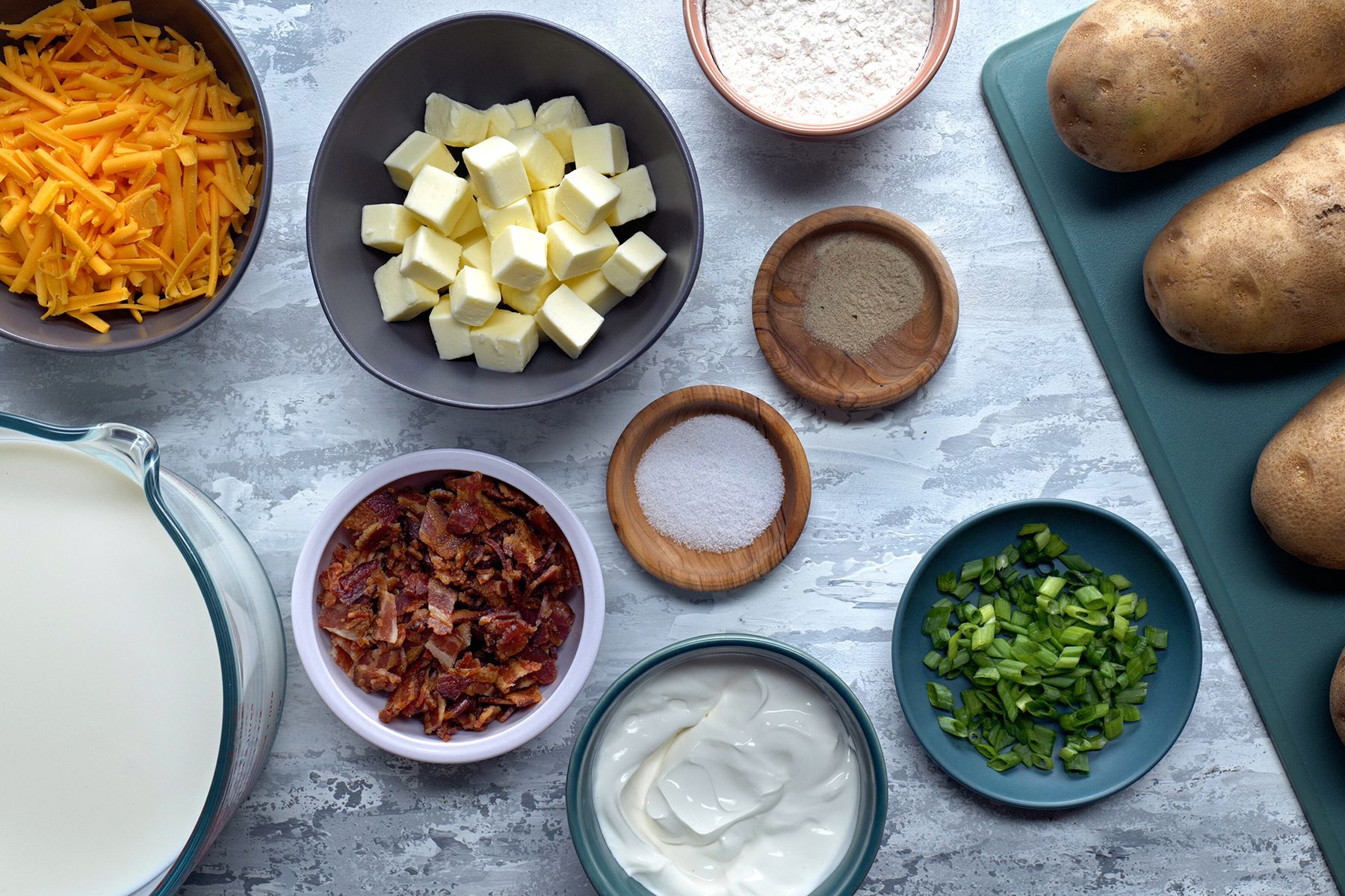 A top-down view of various ingredients arranged on a kitchen counter, including shredded cheese, cubed butter, flour, potatoes, cooked bacon bits, salt, pepper, chopped green onions, sour cream, and a pitcher of milk.
