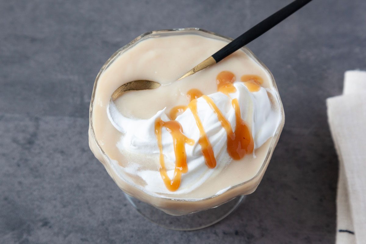 single serving of Taste of Home Caramel Pudding in a glass dessert dish with a small gold and black spoon on a dark gray surface