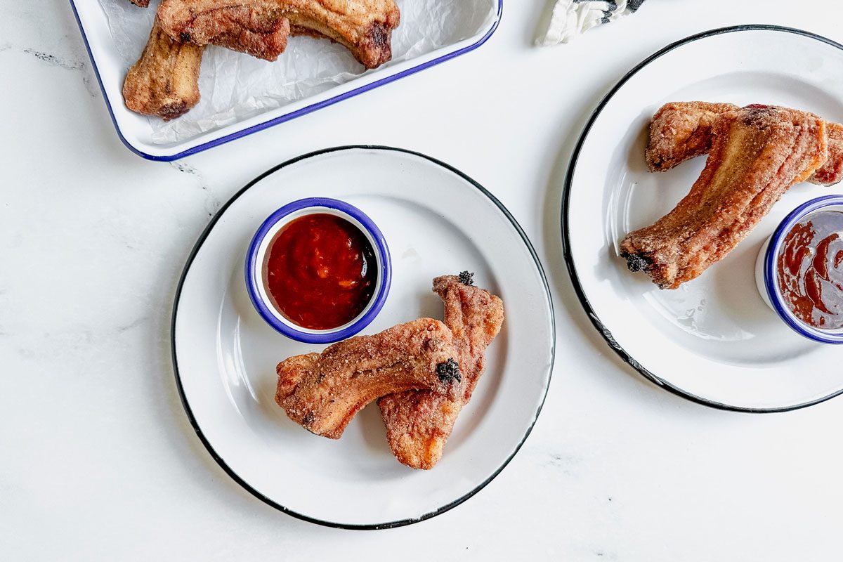 Taste of Home fried ribs on enamel trays and plates with barbecue sauce on a marbe surface next to a cotton napkin.