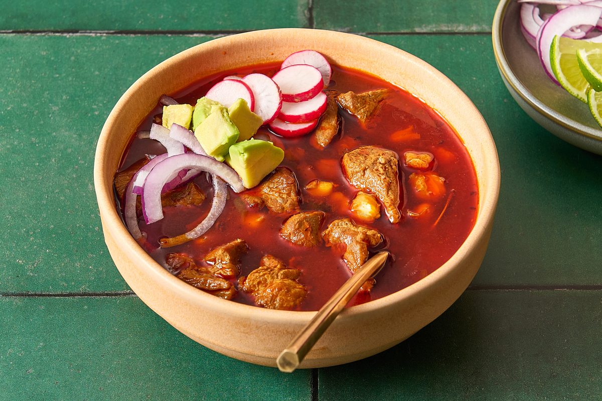 Closeup of a bowl of posole topped with red onion, avocado and radish
