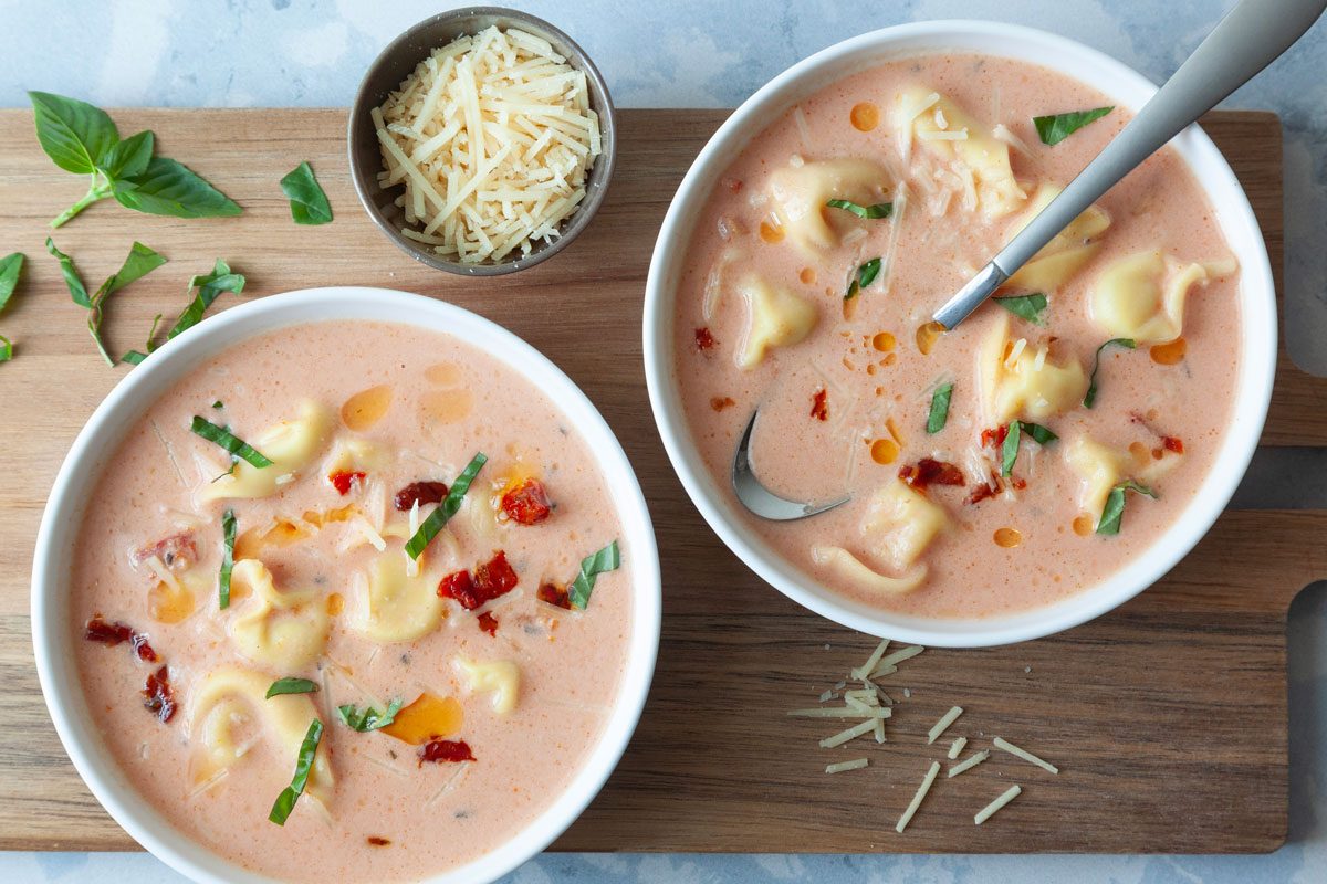 Two single servings of Taste of Home Tomato Tortellini Soup on a wooden cutting board, extra basil and parmesan on board