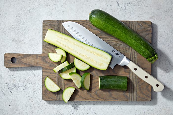 A wooden cutting board displays a partially sliced zucchini, a whole zucchini, and a chopping knife with a white handle.