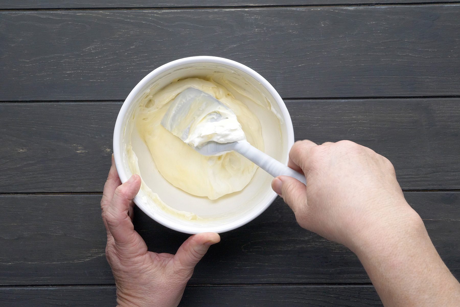 A person stirs a creamy mixture with a white spatula in a white bowl, set on a dark wooden surface. The person's left hand is holding the bowl while the right hand is mixing the creamy contents.