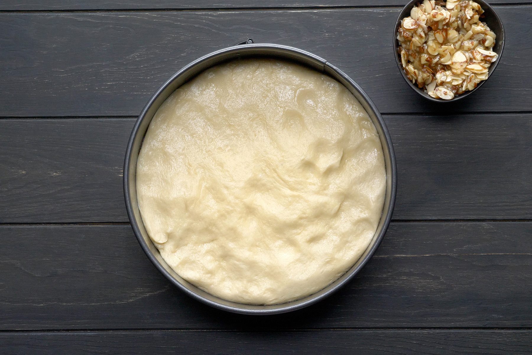 A round baking pan filled with uncooked, spread-out dough sits on a dark wooden surface.