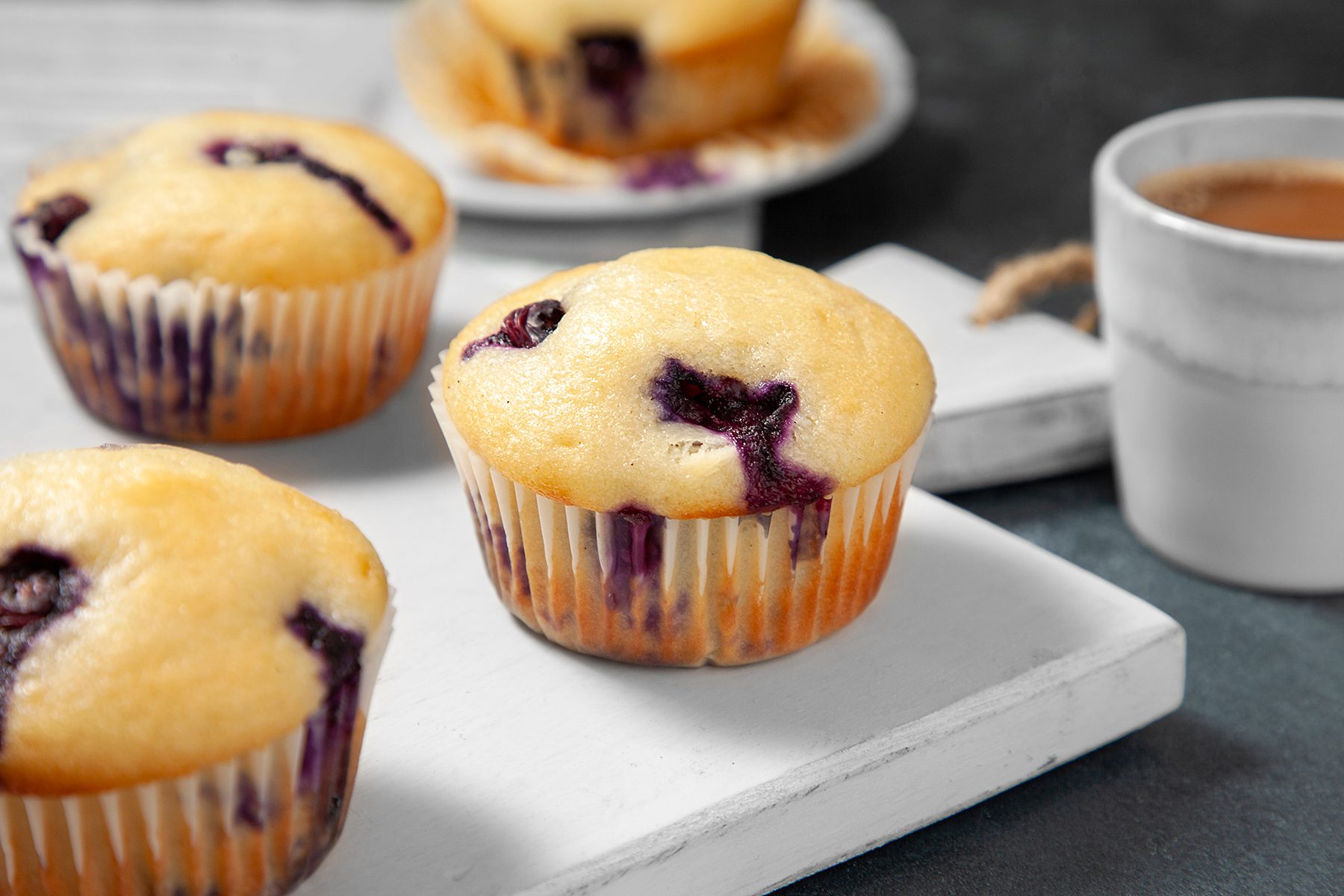 Three blueberry muffins on a white wooden board, with a cup of coffee in the background. Another muffin is partially visible on a small plate in the upper right corner.
