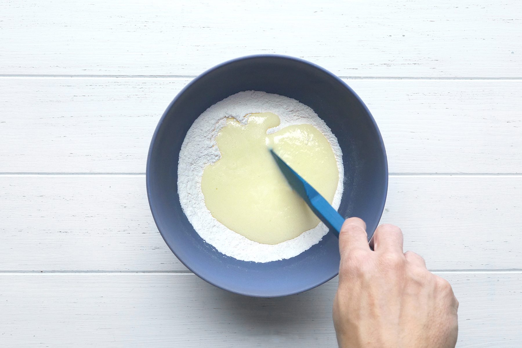 A hand holding a blue spatula mixes wet and dry ingredients in a navy blue bowl.