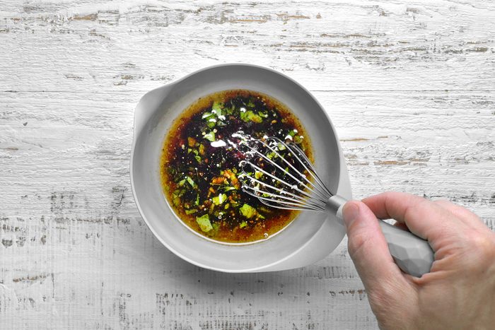 A hand is whisking a dark brown marinade with visible green herbs in a white bowl.