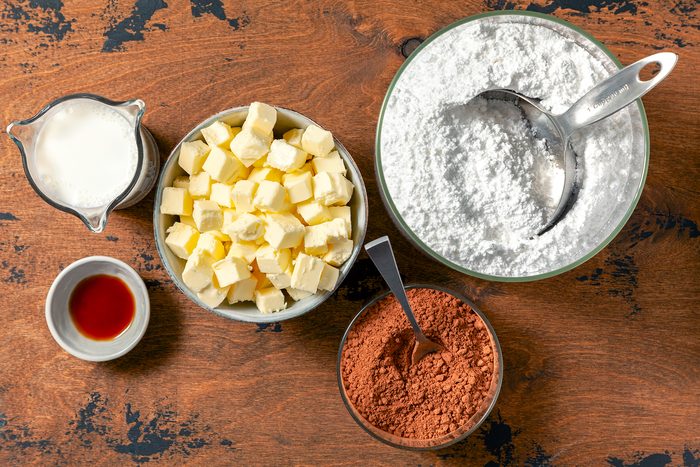 A wooden table with various baking ingredients: a bowl of milk, a bowl of cubed butter, a bowl of powdered sugar with a measuring cup, a bowl of cocoa powder with a spoon, and a small bowl of vanilla extract.