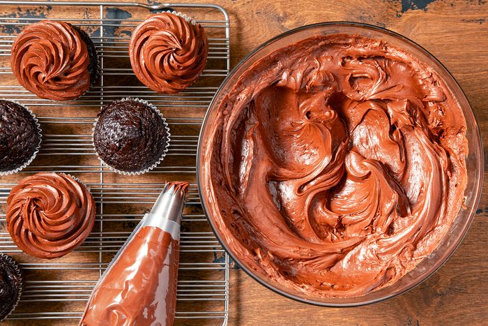 A large bowl filled with smooth, chocolate frosting sits on a wooden surface. Nearby, a wire cooling rack holds several chocolate cupcakes, some of which are already frosted. A piping bag filled with chocolate frosting lies beside the bowl and the cupcakes.