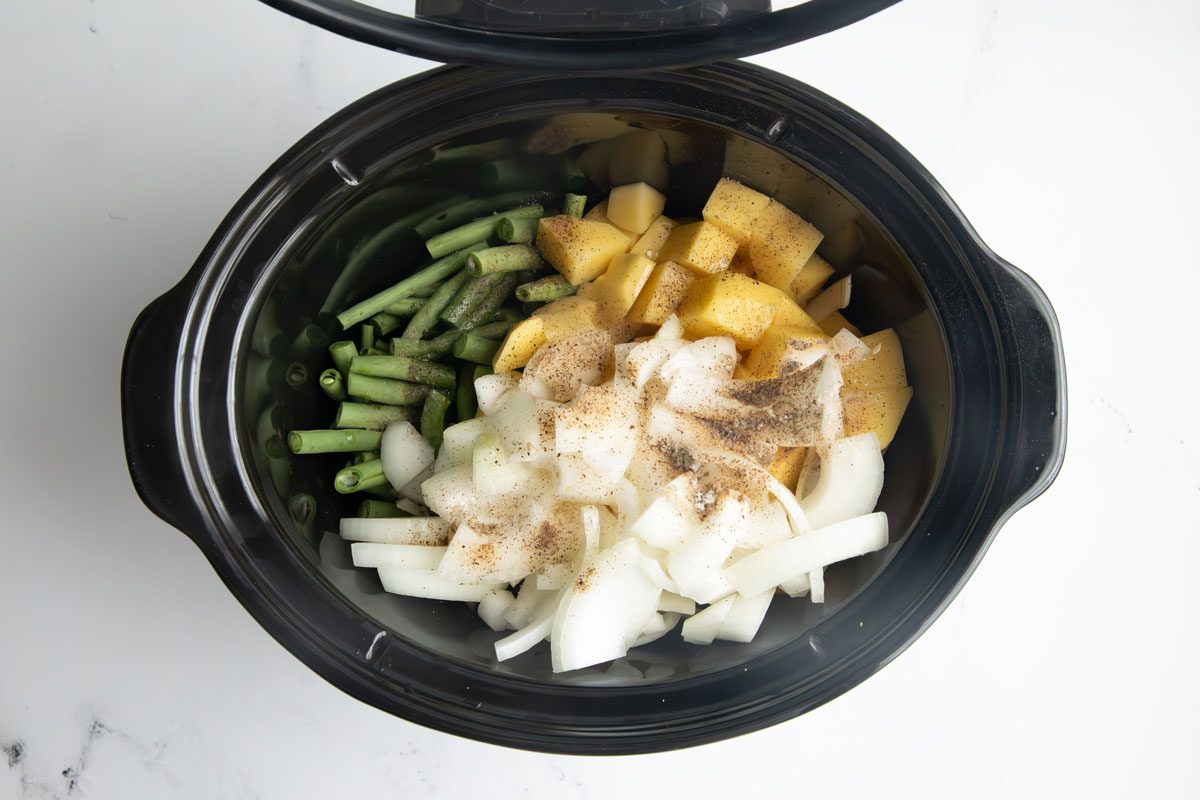 Uncooked green beans, potatoes, onions, salt and pepper in a slow cooker for Taste of home green beans and potatoes on a marble surface.