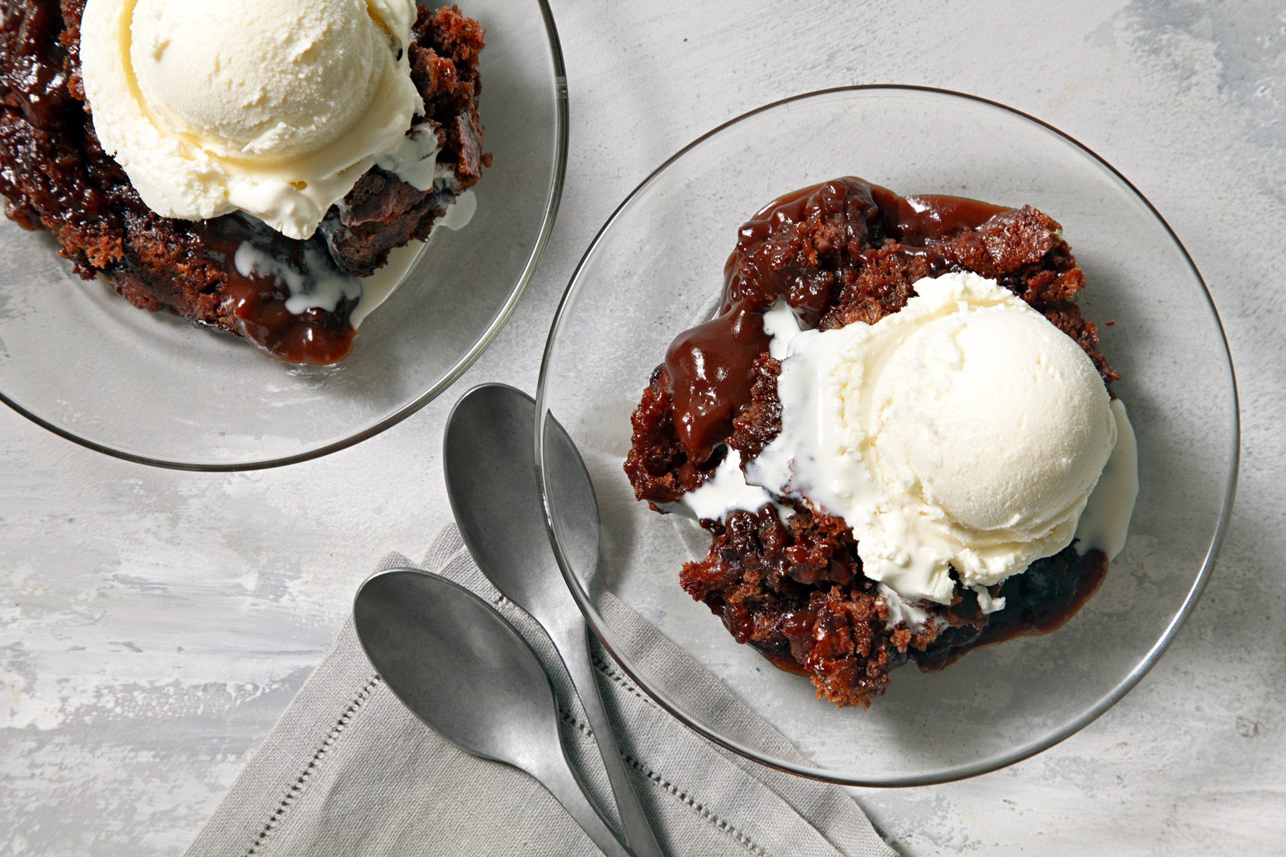 Two glass bowls with hot Fudge Cake with ice cream on top. Two spoons are next to a bowl.