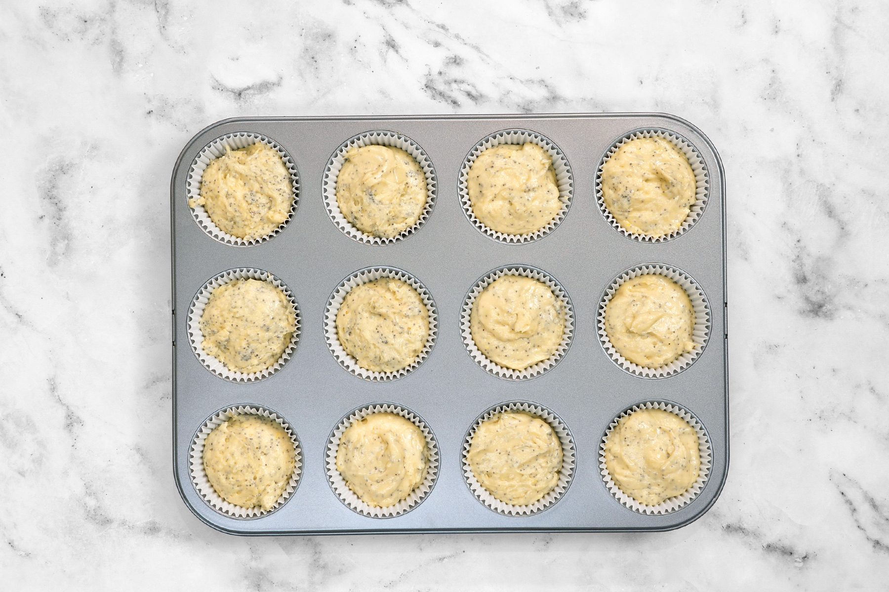 A muffin tin containing 12 portions of uncooked muffin batter, each in a lined cupcake slot, is placed on a white marble countertop.