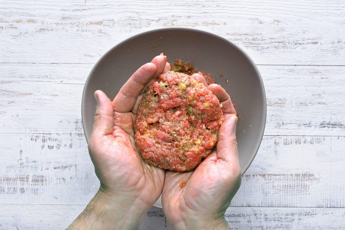 Hands shaping ground meat mixed with spices into a patty over a gray bowl with a rustic white wooden table as the background.