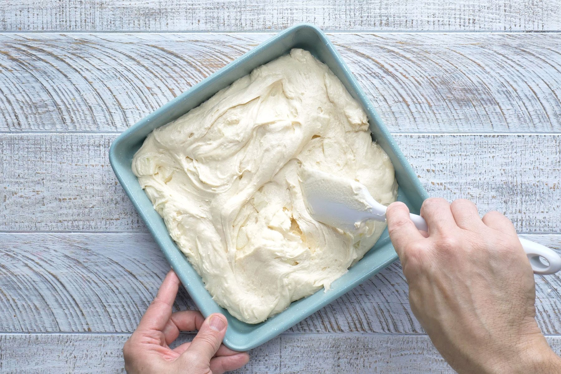 A person's hands can be seen scraping the coffee cake batter into a greased 9-inch square baking pan. The batter is spread evenly to all four corners and edges.