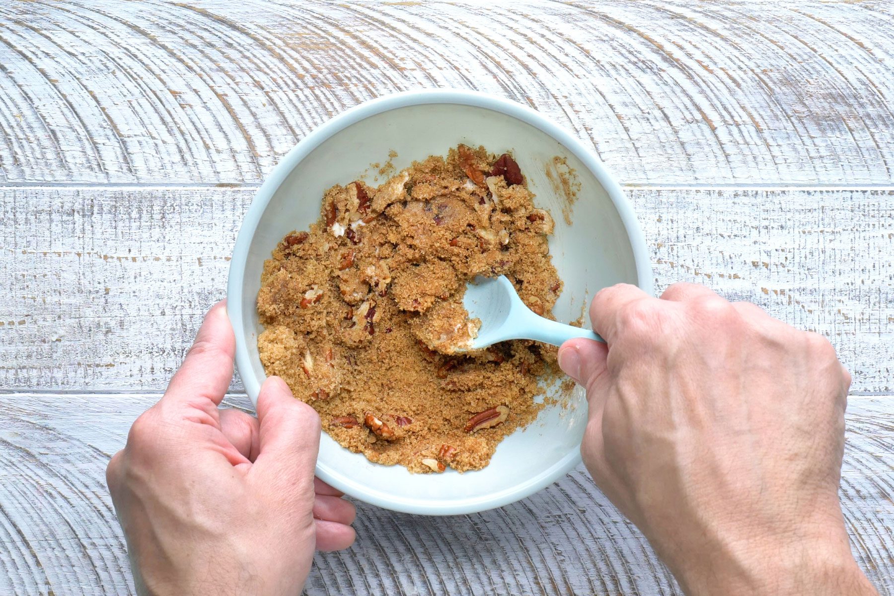 A person's hands are visible stirring together the brown sugar, pecans, softened butter and apple pie spice in a bowl.