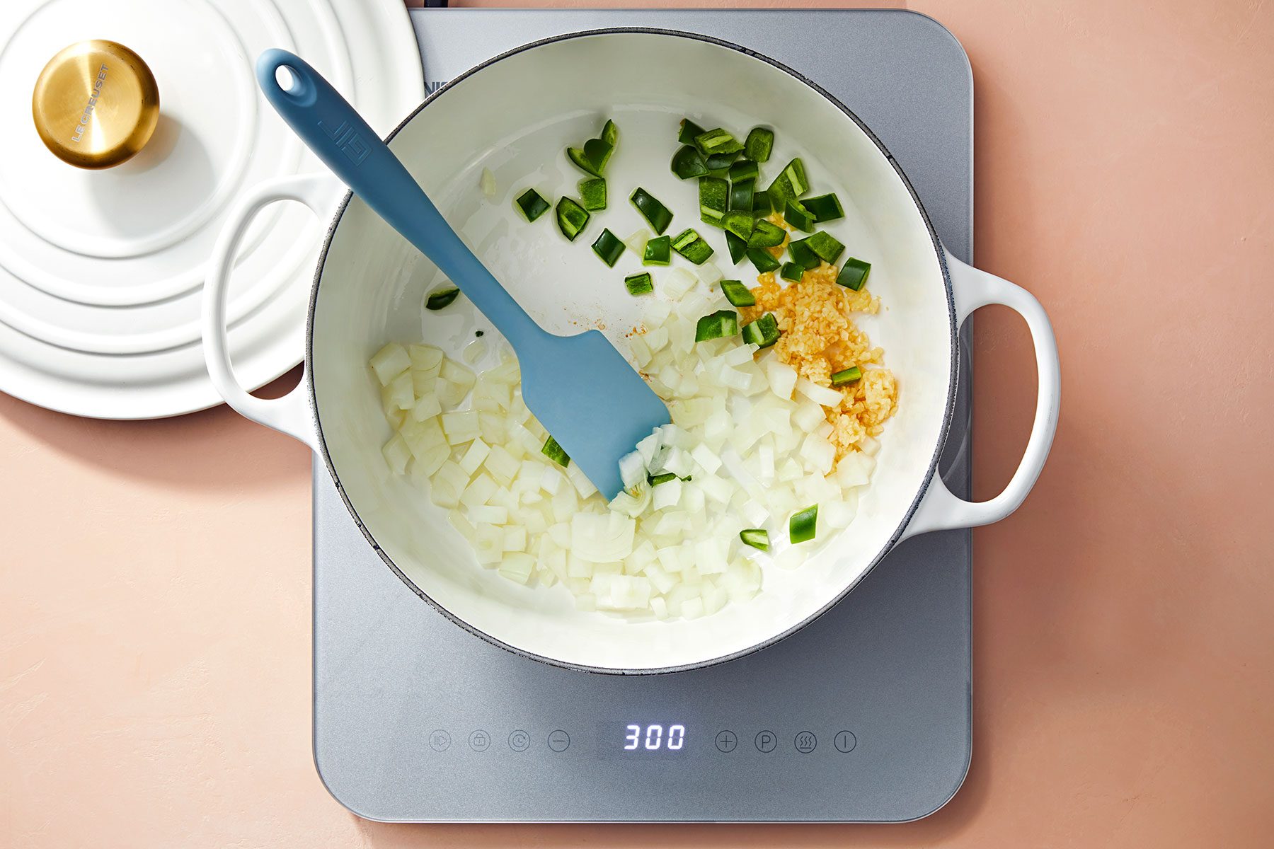 A large white pot on an electric stovetop contains chopped onions, garlic, and green peppers being stirred with a blue spatula. A white lid with a gold handle is partially visible in the background. The stovetop display shows "300.