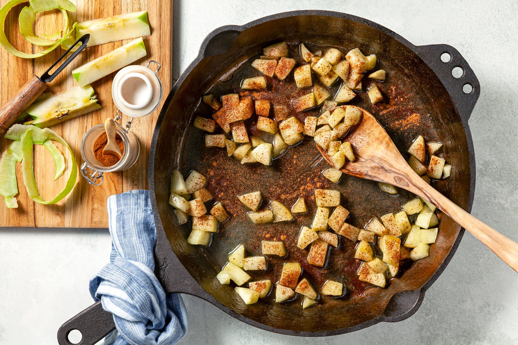 overhead shot; white background; a cast-iron skillet filled with diced apples, being sautéed in butter; The apples are a light brown color and are coated in a mixture of spices, including cinnamon and sugar; There is a wooden cutting board with apple peels and a small bowl of cinnamon on the side;