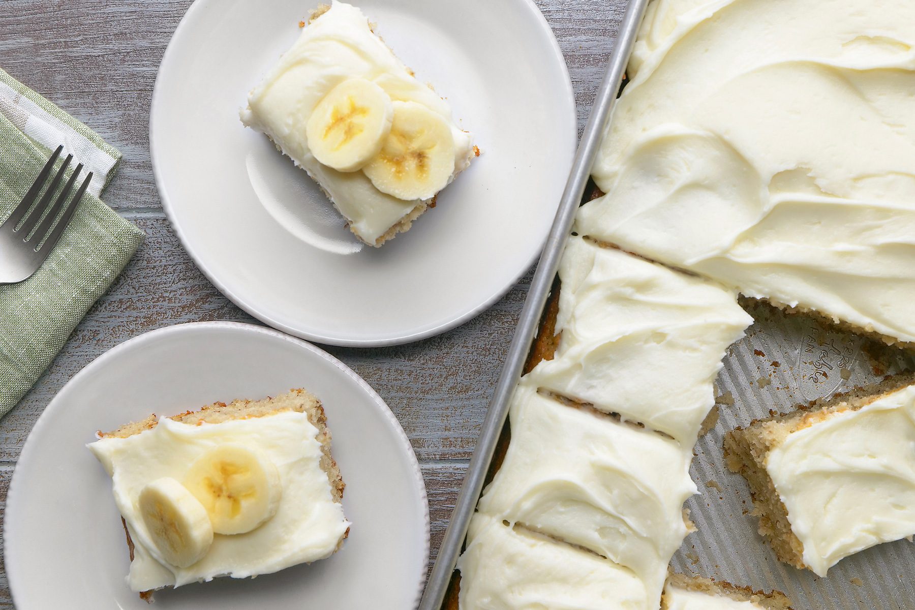 overhead shot, grey background, A delicious-looking banana cake is featured in the image, A rectangular baking sheet holds a whole, frosted banana cake, while two smaller plates each contain a slice of the cake topped with banana slices; The cake has a golden-brown crust and is covered in a creamy white frosting