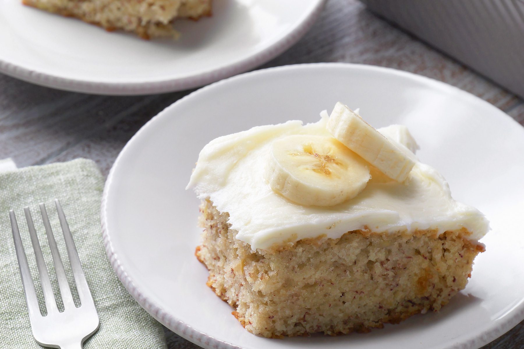 3/4th shot, grey background, a delightful slice of banana cake, perfectly frosted with a creamy white icing and topped with fresh banana slices; A fork rests on a green napkin