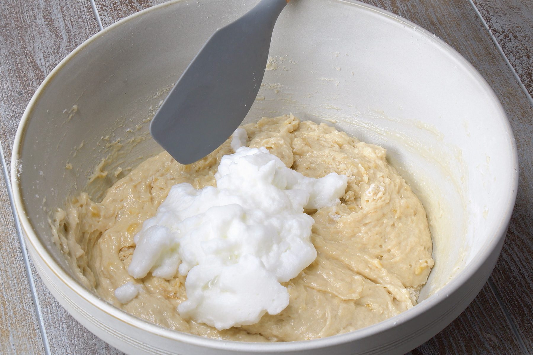 3/4th shot; grey background; A white bowl contains a batter with a light brown color, Mounds of whipped egg whites are being gently folded into the batter using a spatula; The egg whites are white and fluffy, contrasting with the darker batter