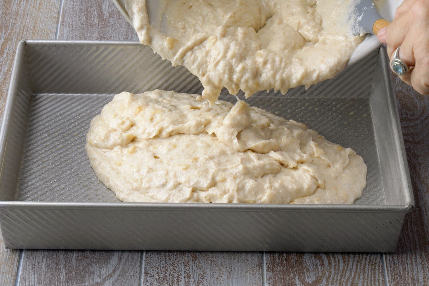 3/4th shot; grey background; A batter is being poured into a baking pan, The batter is a light brown color and has a thick, creamy consistency, The baking pan is rectangular and has a dark gray color