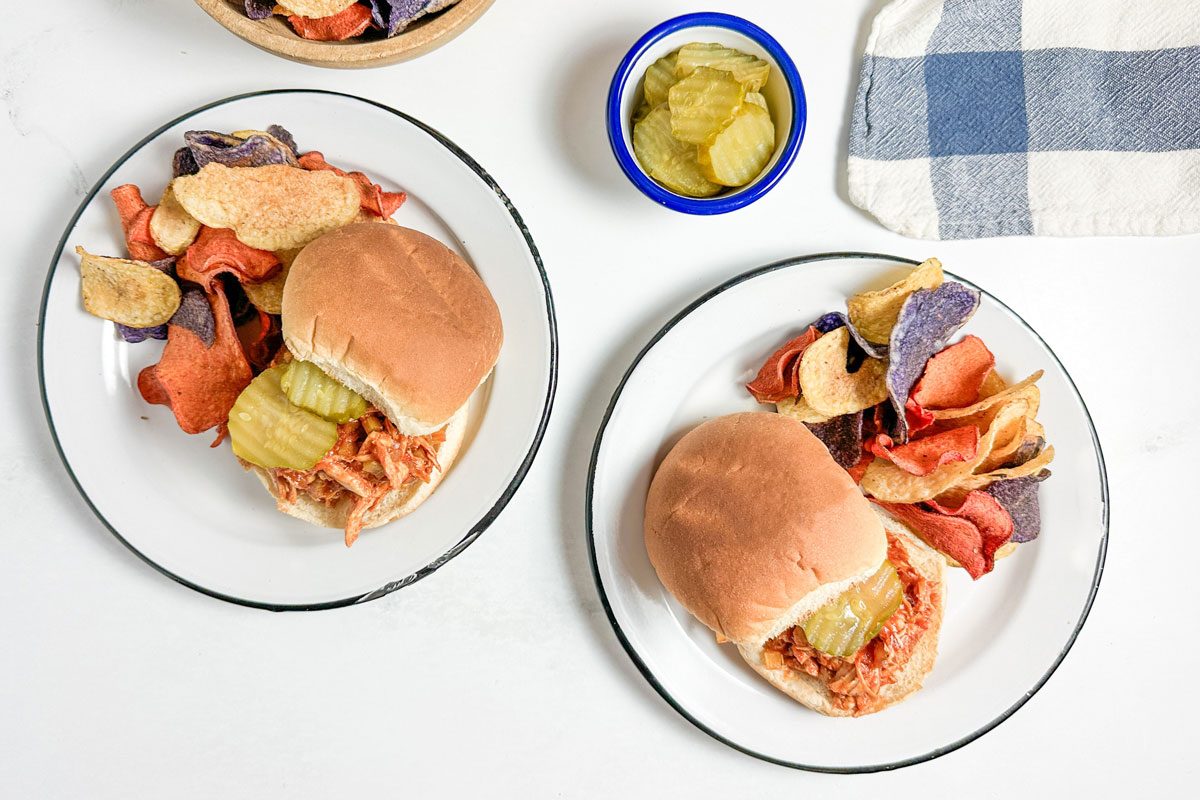 Taste of Home Barbecue Chicken Sandwich with chips on enamel plates next to bowls of chips and pickles