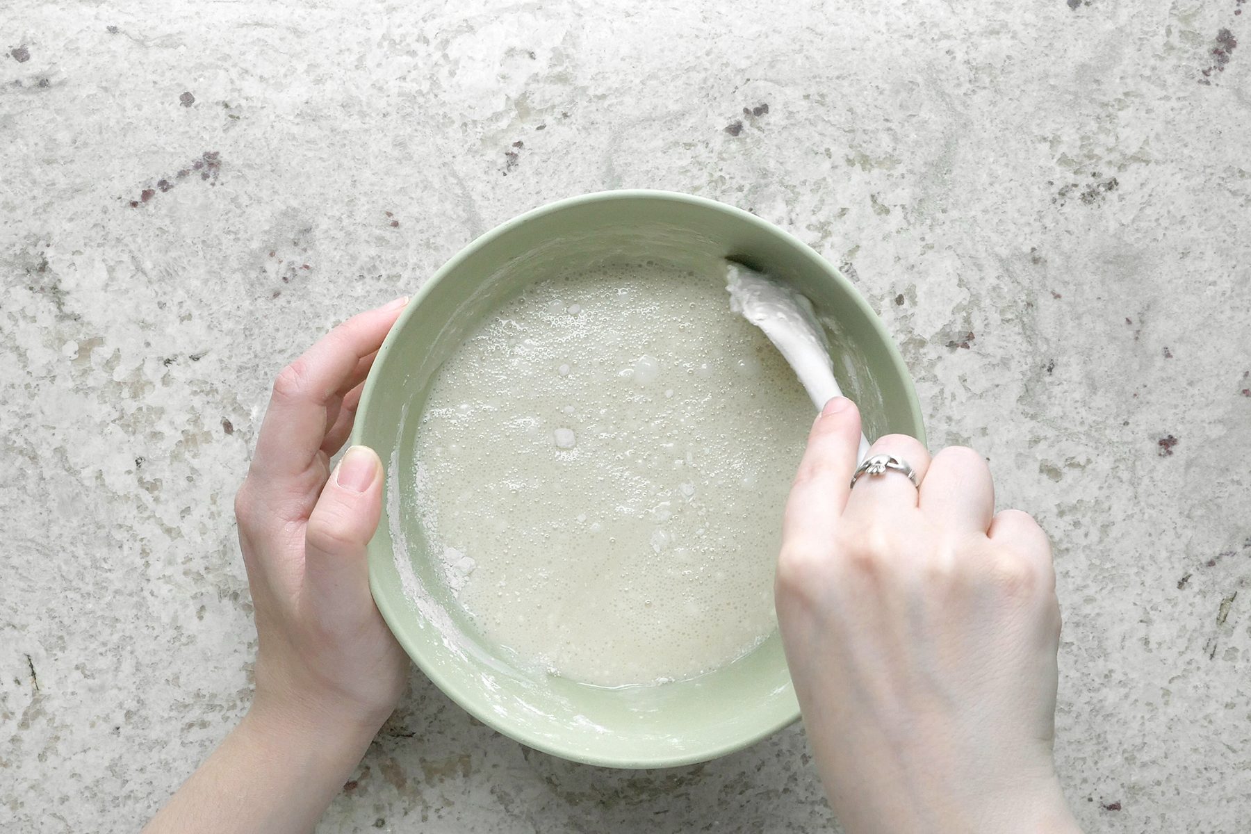 A top view of a person's hands stirring a mixture in a light green bowl with a white spatula. The mixture appears wet and frothy. The background is a light, speckled countertop. The person is wearing a ring on their left hand.