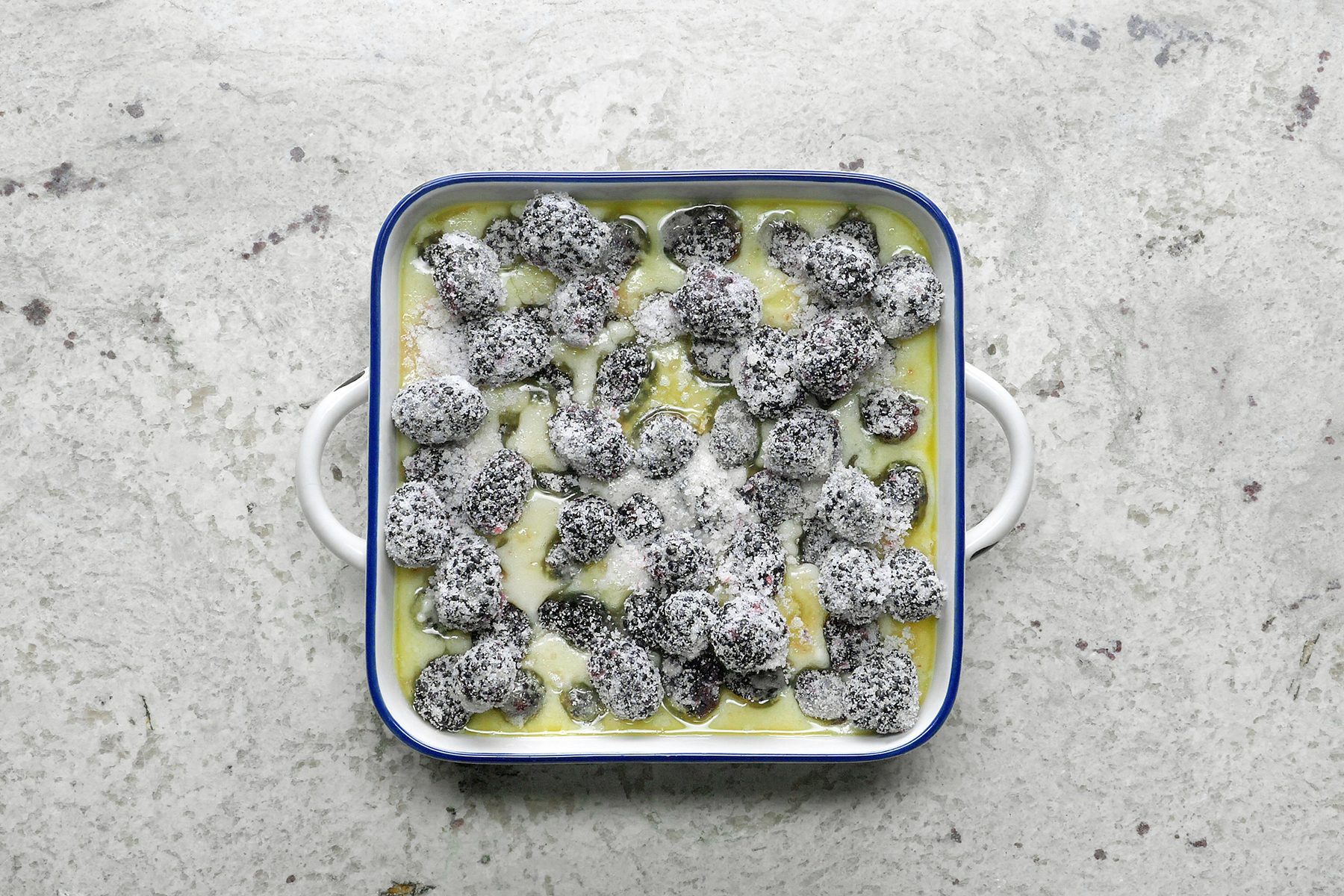 Top view of a white baking dish with blue trim filled with what appears to be a creamy baked dish, possibly featuring coated or dusted small dark objects, on a textured, light-colored surface.