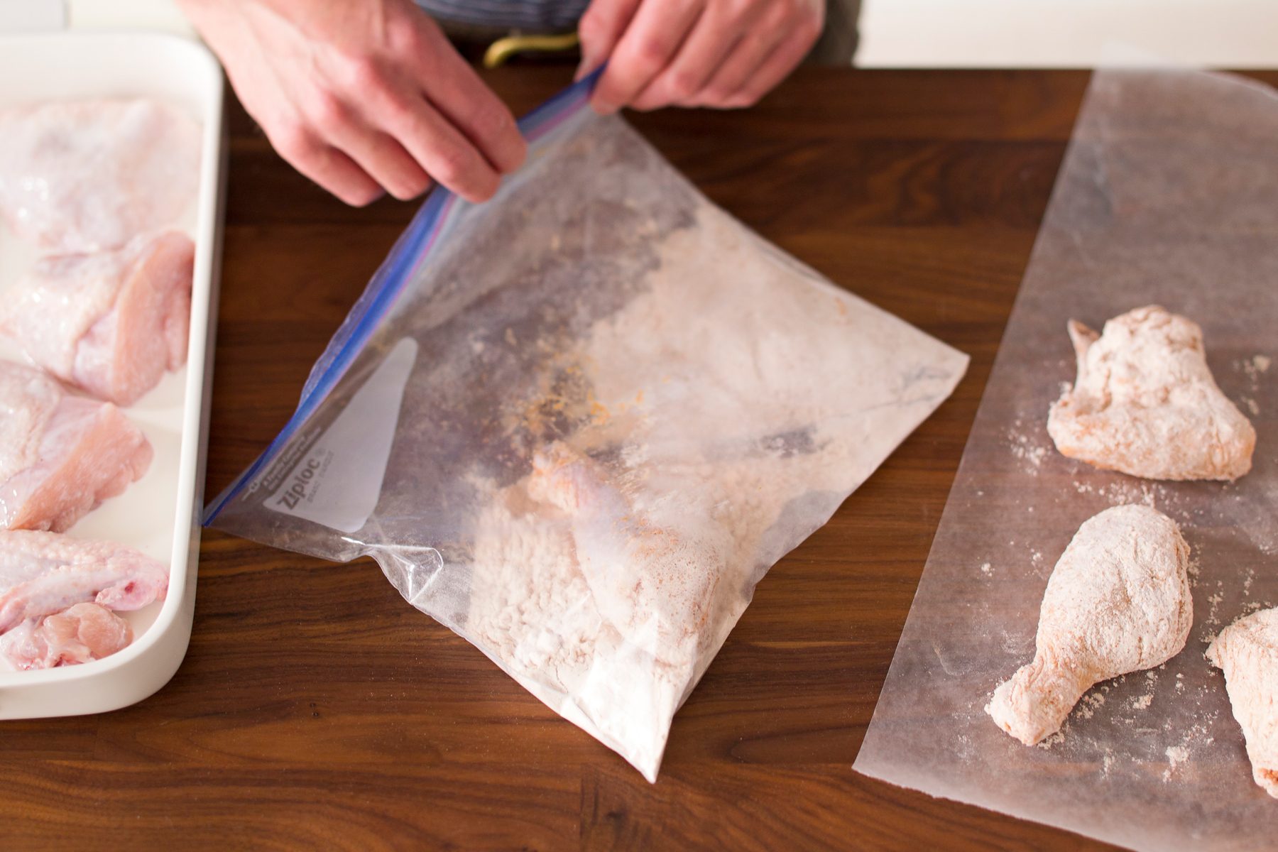 A person taking a drumstick from its buttermilk bath and getting it covered in coating inside a large resealable plastic bag then placing it on parchment paper to their left