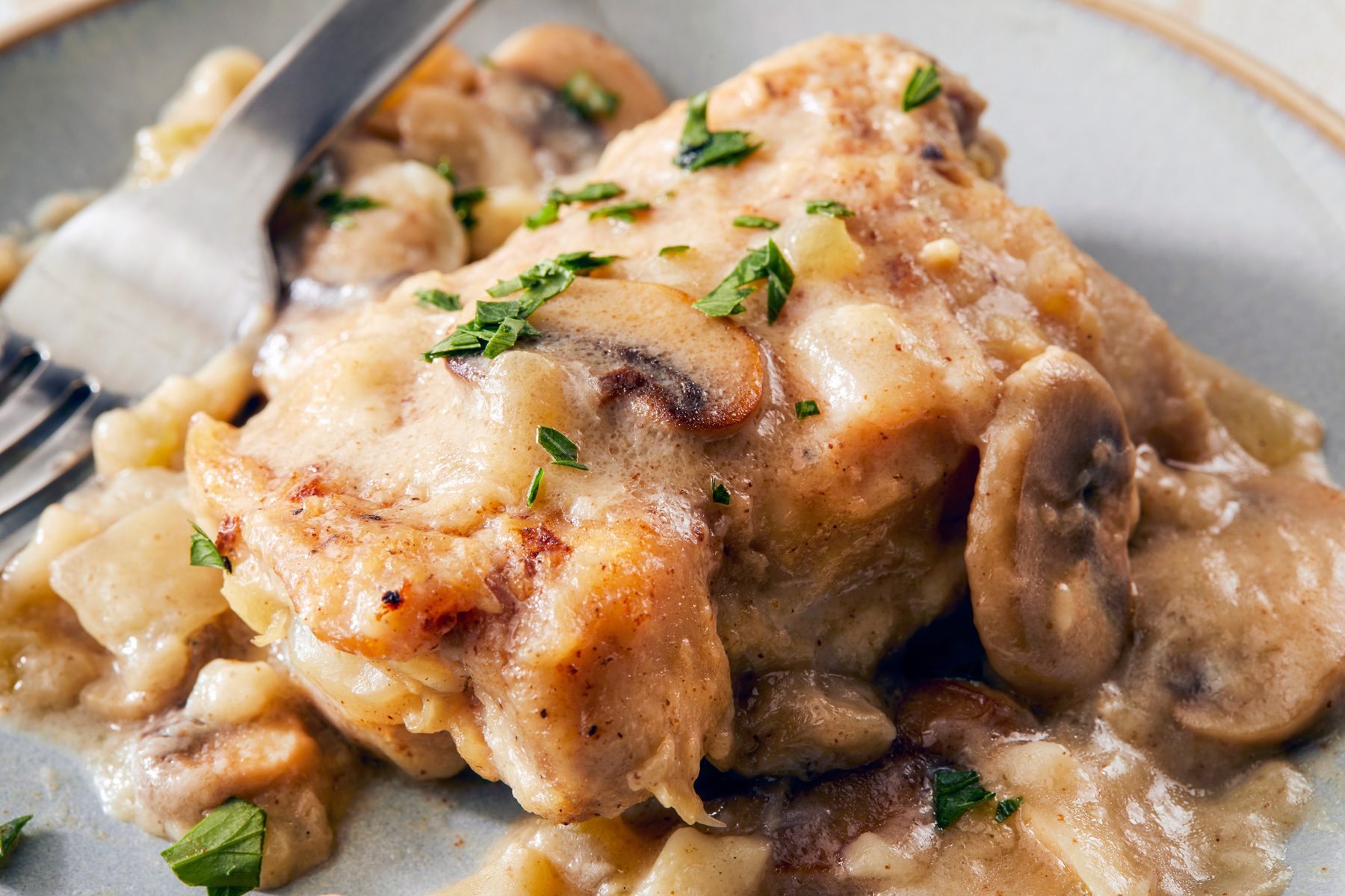 close shot; A plate of Chicken Fricassee is presented, featuring tender chicken thighs smothered in a creamy, mushroom-rich sauce; The sauce is a pale golden color, and the mushrooms are visible throughout; Fresh parsley is sprinkled on top