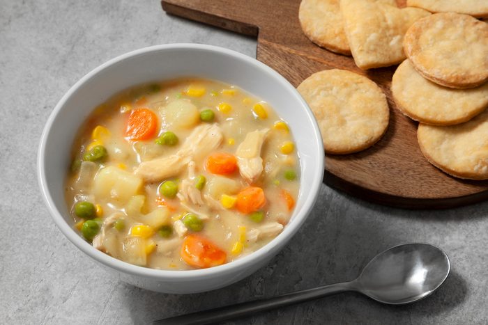 3/4th shot; light grey background; A bowl of Chicken Pot Pie Soup is featured, filled with tender chicken, vegetables, and a creamy broth; Heart-shaped biscuits are arranged on a wooden cutting board