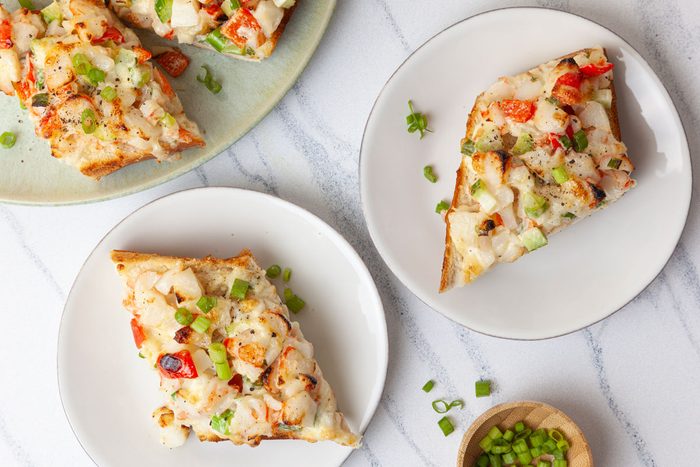 Single servings of Taste of Home Crab Salad Sandwiches on white app plates, full yield platter peaking in.