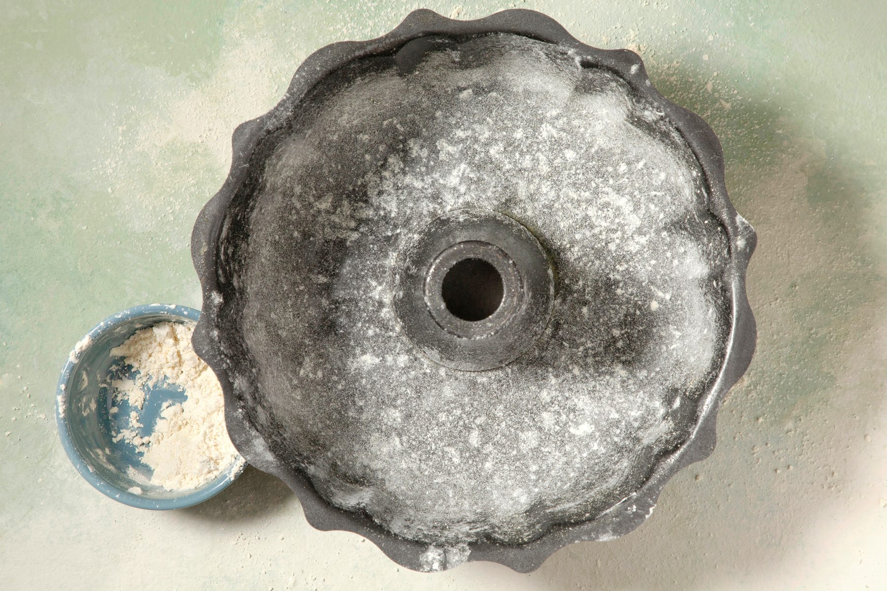 overhead shot; light green textured background; Grease and flour a fluted tube pan using gluten-free flour; flour in small bowl next to fluted tube pan