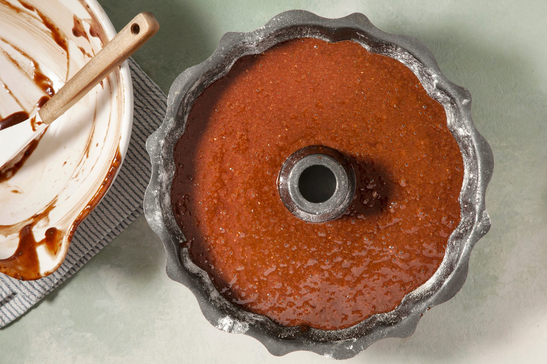 overhead shot; light green textured background; A dark chocolate cake batter is poured into a fluted bundt pan; The batter fills the pan almost to the top, A small bowl with a spatula and some batter residue is placed next to the pan
