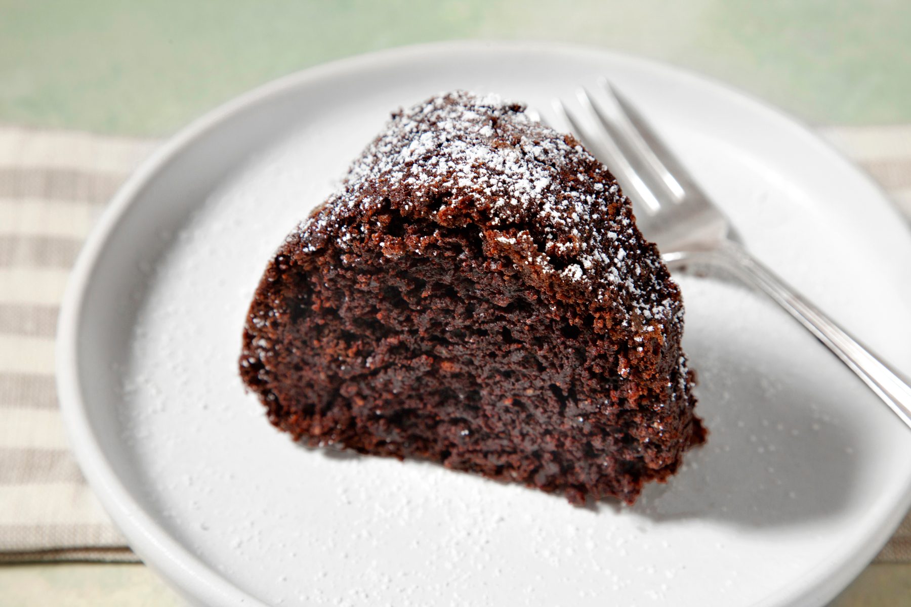 close shot; light green background; A slice of chocolate cake on a white plate; It's dusted with powdered sugar; silver fork placed on white plate over kitchen towel