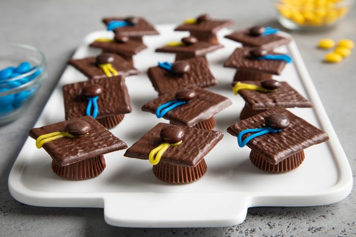 A white tray holds a dozen graduation cap-themed chocolate treats. Each treat is composed of an upside-down chocolate peanut butter cup topped with a square of chocolate, adorned with a candy-coated chocolate and a colorful frosting tassel.