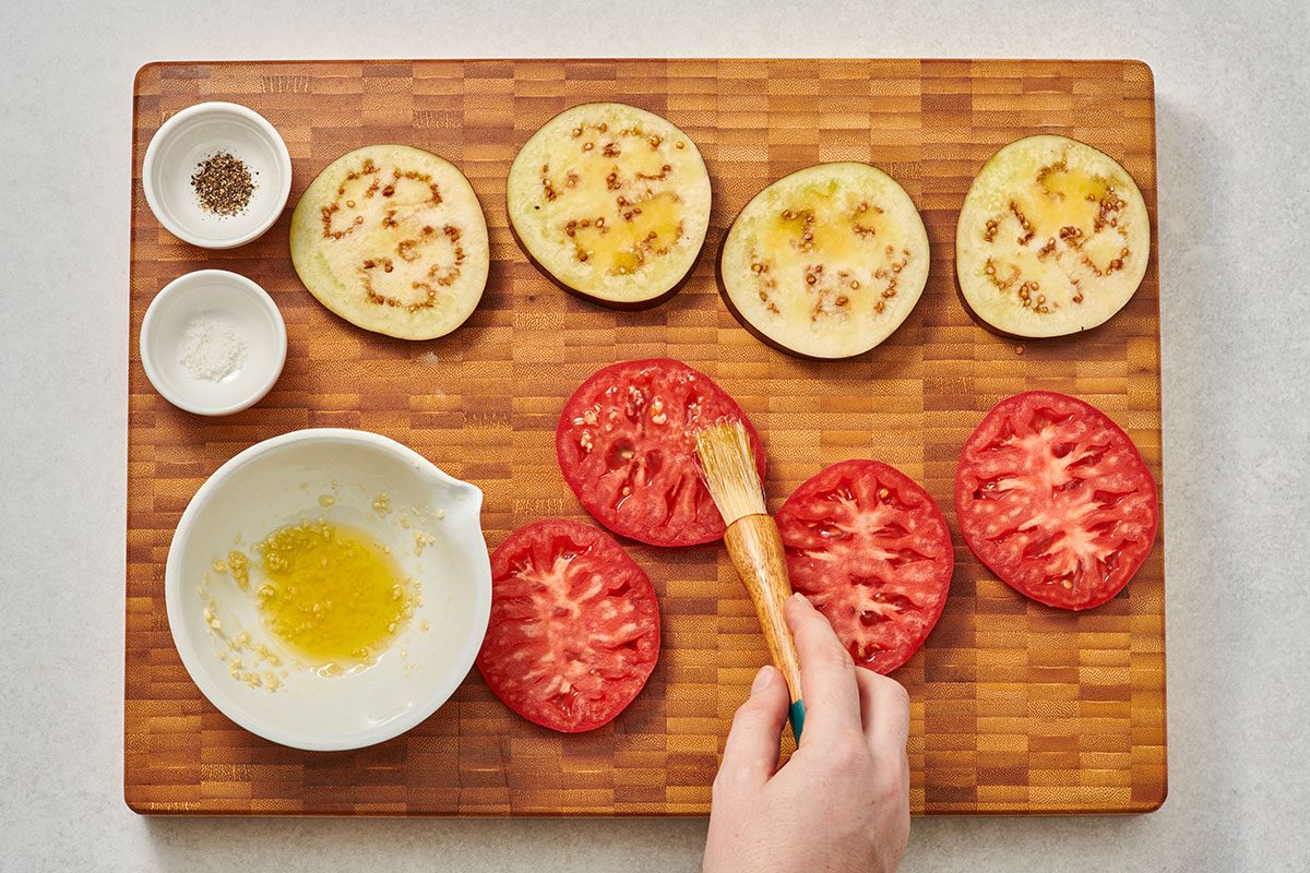 Brushing tomato and eggplant slices with olive oil and garlic paste on a cutting board