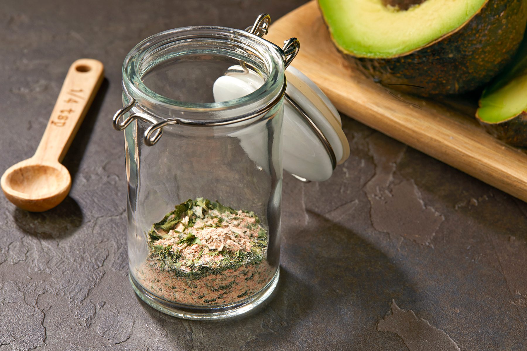 A glass jar containing a blend of spices and herbs sits open on a dark textured surface. Beside it, there's a wooden measuring spoon marked "1/4 tsp," and a halved avocado on a wooden cutting board is partially visible in the background.