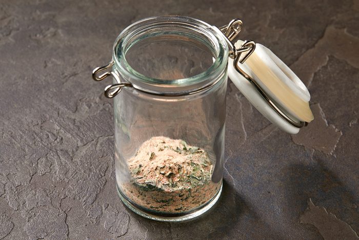 A clear glass jar with a hinged white lid contains a small amount of mixed powdered substance in various shades of brown, beige, and green. The jar stands on a textured, dark-colored surface.