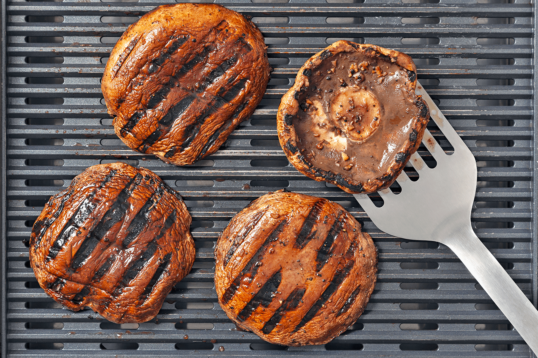 Four grilled portobello mushroom caps with char marks are on a grill pan. A metal spatula holds one of the mushrooms up, exposing the gills and underside of the cap. The mushrooms are evenly browned and appear juicy.