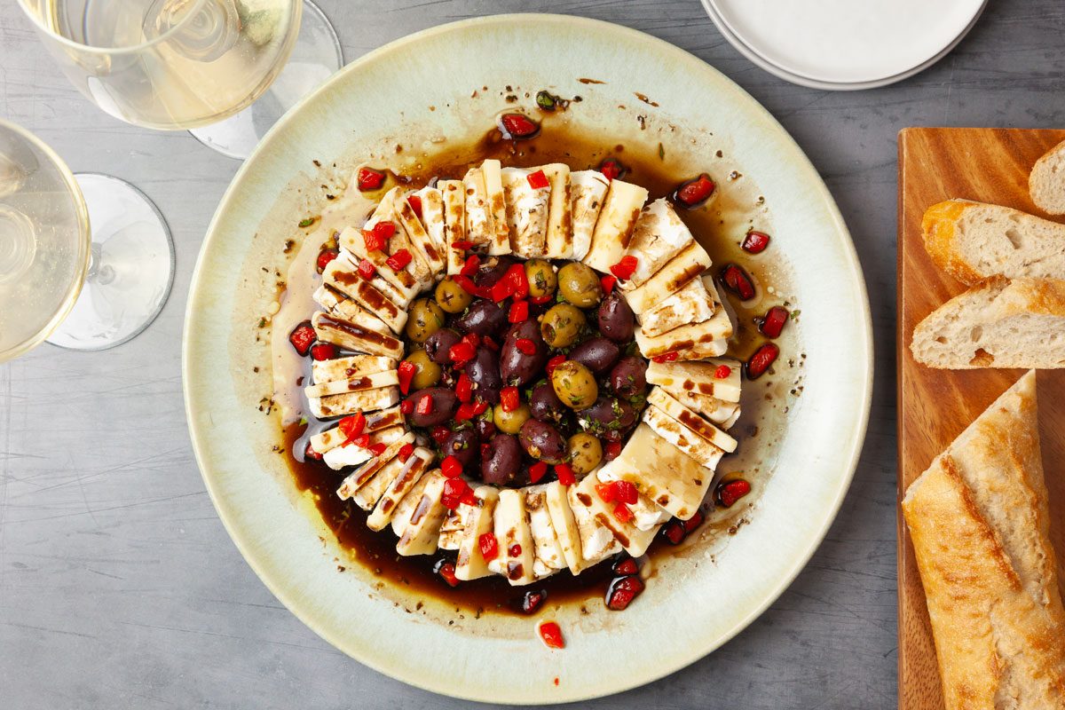 Full yield of Taste of Home Olives & Cheese on a serving plate, sliced baguette on a cutting board, white wine