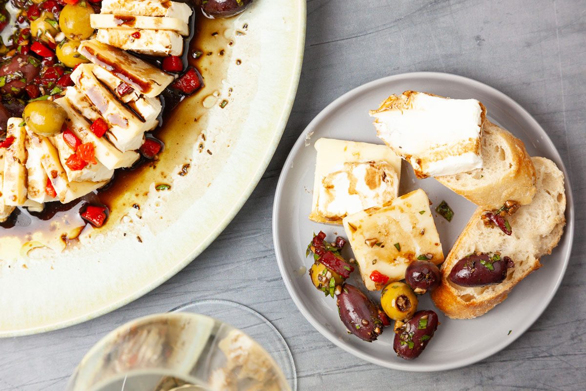 Close up of single servings of Taste of Home Olives & Cheese and full yield peakin in on a serving plate, sliced baguette on a cutting board, white wine