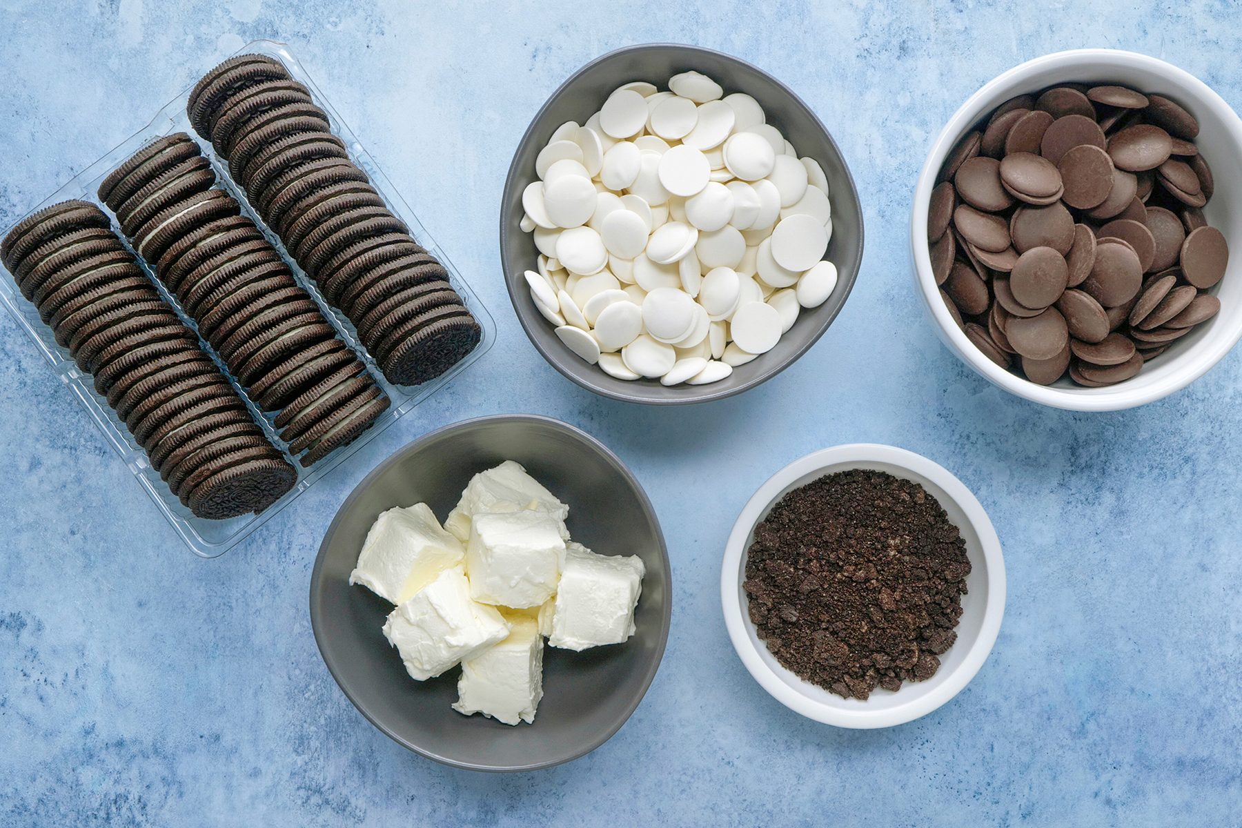 Ingredients for making chocolate truffles are arranged on a blue surface, including Oreo cookies in a packaging, white chocolate chips, milk chocolate discs, cream cheese, and ground Oreo crumbs in small bowls.
