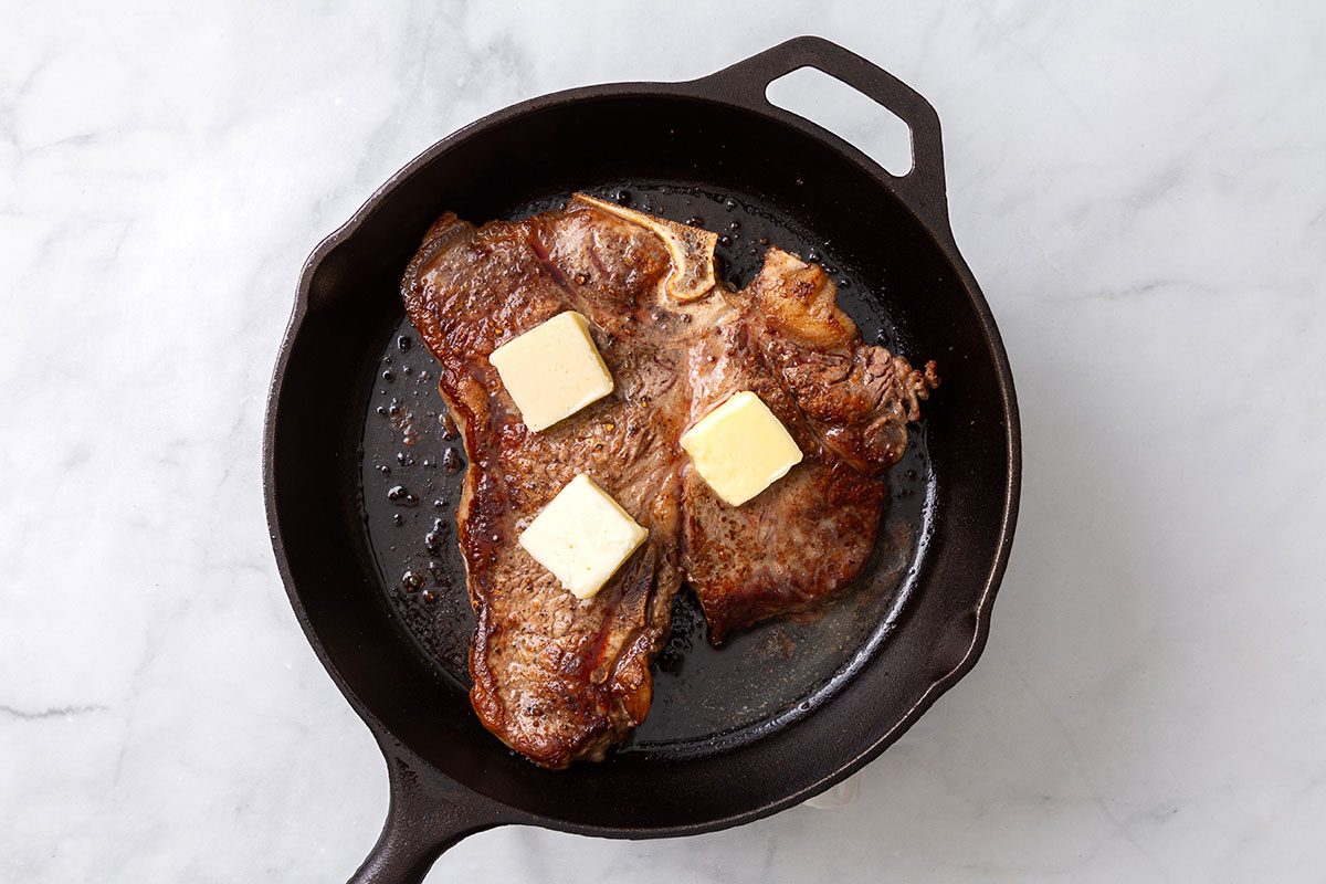 Seared steak topped with butter in a cast iron skillet before being put in the oven for step three for Porterhouse Steak recipe for Taste of Home